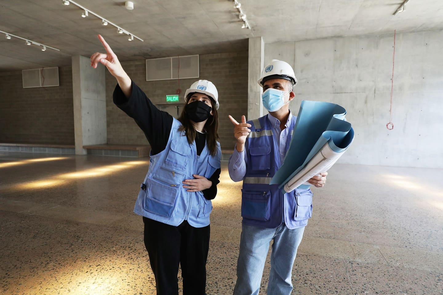 Two UNOPS staff in hard hats and field vests inspecting the interior of a building under construction, one carrying rolled drawings